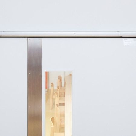 Zwei Doppelbrandschutztüren mit rechteckigem Fenster in der rechten Tür. Die Türen haben Türschließer oben und einen Metallstreifen in der Mitte. Durch das Fenster sind Holzstühle zu sehen