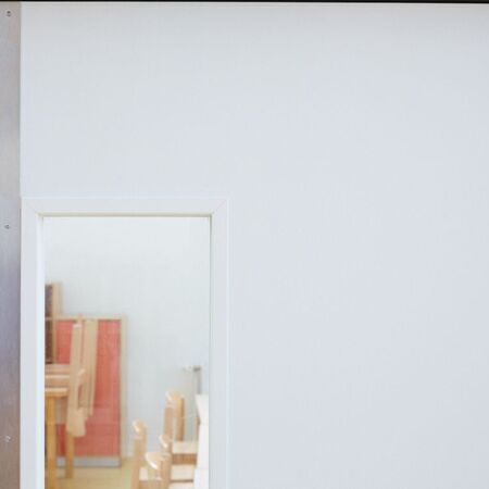 Brandschutztür mit kleinem rechteckigem Fenster, durch das ein Raum mit Holzstühlen und Tischen zu sehen ist. Links an der Wand befindet sich ein Metallstreifen. Oben rechts im Bild ist ein kleines Etikett mit Text