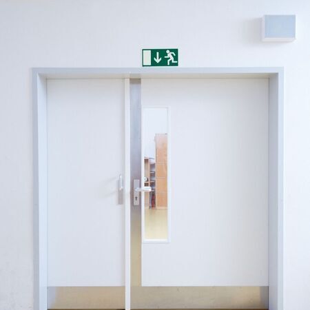 Doppelbrandschutztüren mit rechteckigem Fenster und grünem Notausgangsschild darüber. Metallgriffe und heller Flur