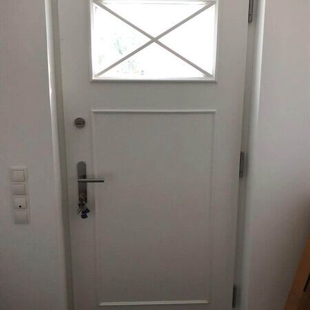 Haustür im Landhausstil mit rechteckigem Fenster oben. Silberner Türgriff mit Schlüsseln. Links drei Lichtschalter an der Wand