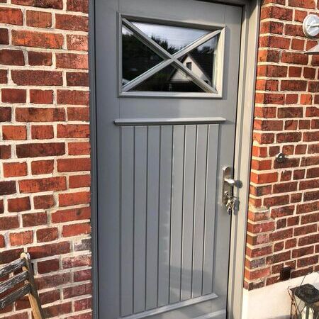 Graue Haustüre zweifarbig im Landhausstil in einer roten Ziegelwand mit rechteckigem Fenster oben. Silberner Griff mit Schlüsseln. Rechts eine Außenleuchte und ein Türknopf. Vor der Tür ein kleiner Metallstuhl und eine Laterne