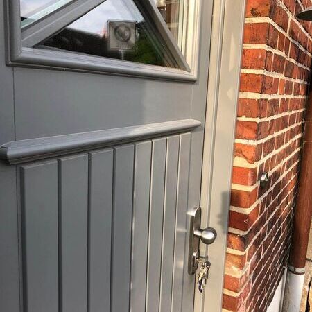 Graue Haustür im Landhausstil mit geometrischem Design und Glasfenster oben. Silberner Griff mit Schlüsseln im Schloss. Tür in einer Ziegelwand mit Abflussrohr rechts