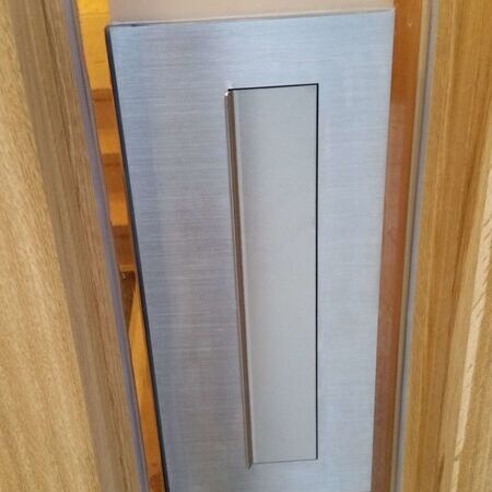 Detailaufnahme eines modernen Briefkastenschlitzes in einer Holz- und Glasschiebetür, mit einem Edelstahlabdeckblech, das leicht geöffnet wird
