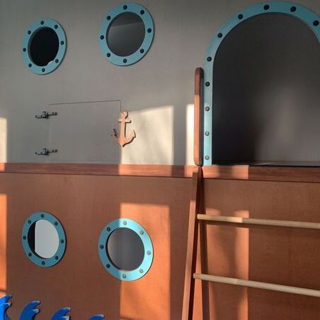 Spielebene in der Kita mit einer Wand in Schiffsoptik, runden Bullaugen-Fenstern und einer Holzleiter, ergänzt durch maritime Details wie eine Anker-Dekoration