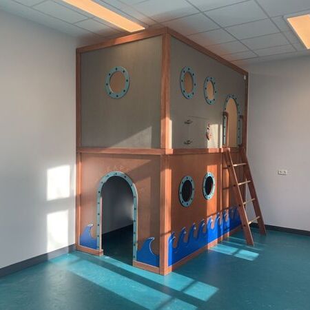 Spielebene in der Kita mit einer Wand in Schiffsoptik, runden Bullaugen-Fenstern und einer Holzleiter, ergänzt durch maritime Details wie eine Anker-Dekoration