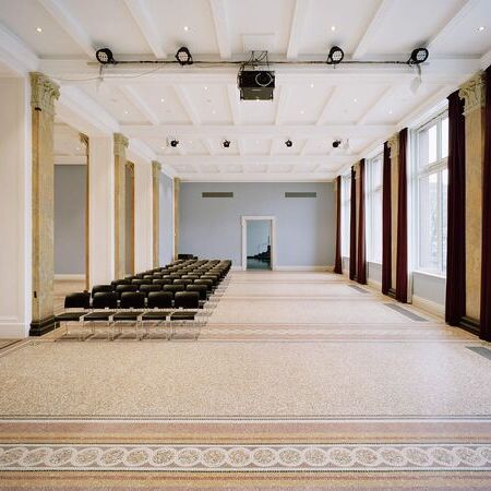 Großer Raum mit eleganten Säulen, einem Mosaikboden und mehreren Stuhlreihen, die auf eine Wand mit großen Fenstern und roten Vorhängen ausgerichtet sind. Über dem Raum verlaufen Scheinwerfer und Lautsprecher an der Decke