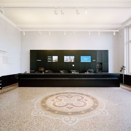 Moderne Empfangstheke in der Kunsthalle Hamburg mit elegantem Mosaikboden und einer Wand mit Informationsschildern. Die Theke ist mit schwarzen Regalen und Bildschirmen ausgestattet, und es gibt eine Vielzahl an Broschüren auf der linken Seite
