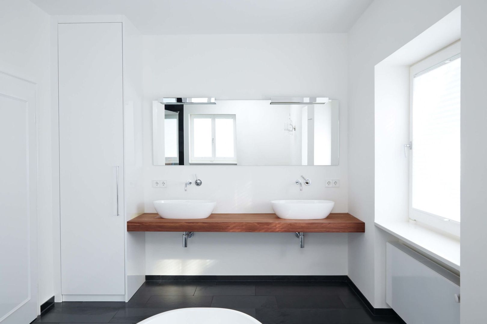 Minimalistisches Badezimmer mit zwei weißen Aufsatzwaschbecken auf einer Holzplatte, großer Spiegel darüber und einem Fenster im Hintergrund