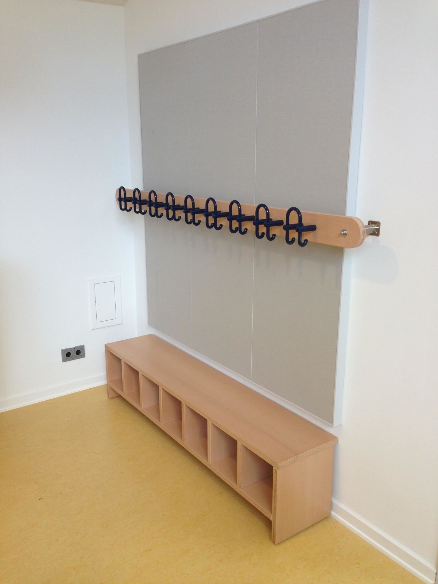 Garderobe in Kita - Holzbank mit sechs Fächern darunter. Darüber ein Holzregal mit Haken. Gelber Bodenbelag