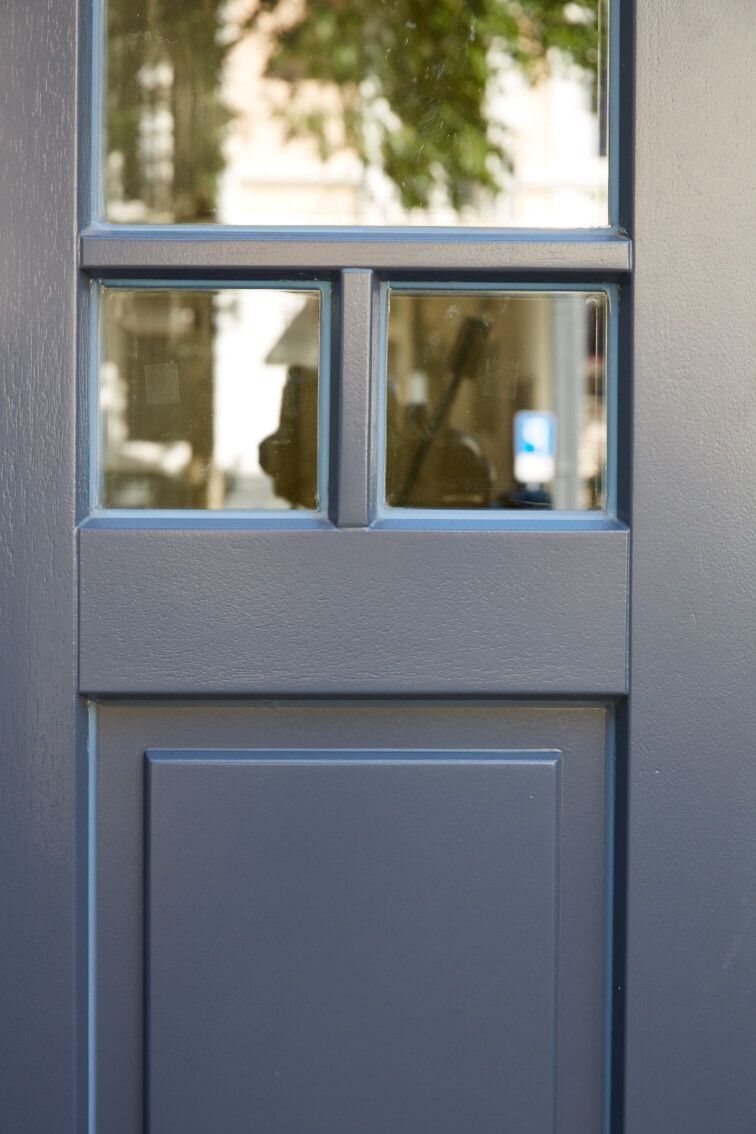 Nahaufnahme einer grauen Tür mit quadratischen Glaseinsätzen und einem Türgriff in Edelstahl, wobei das reflektierte Licht der Umgebung sichtbar ist