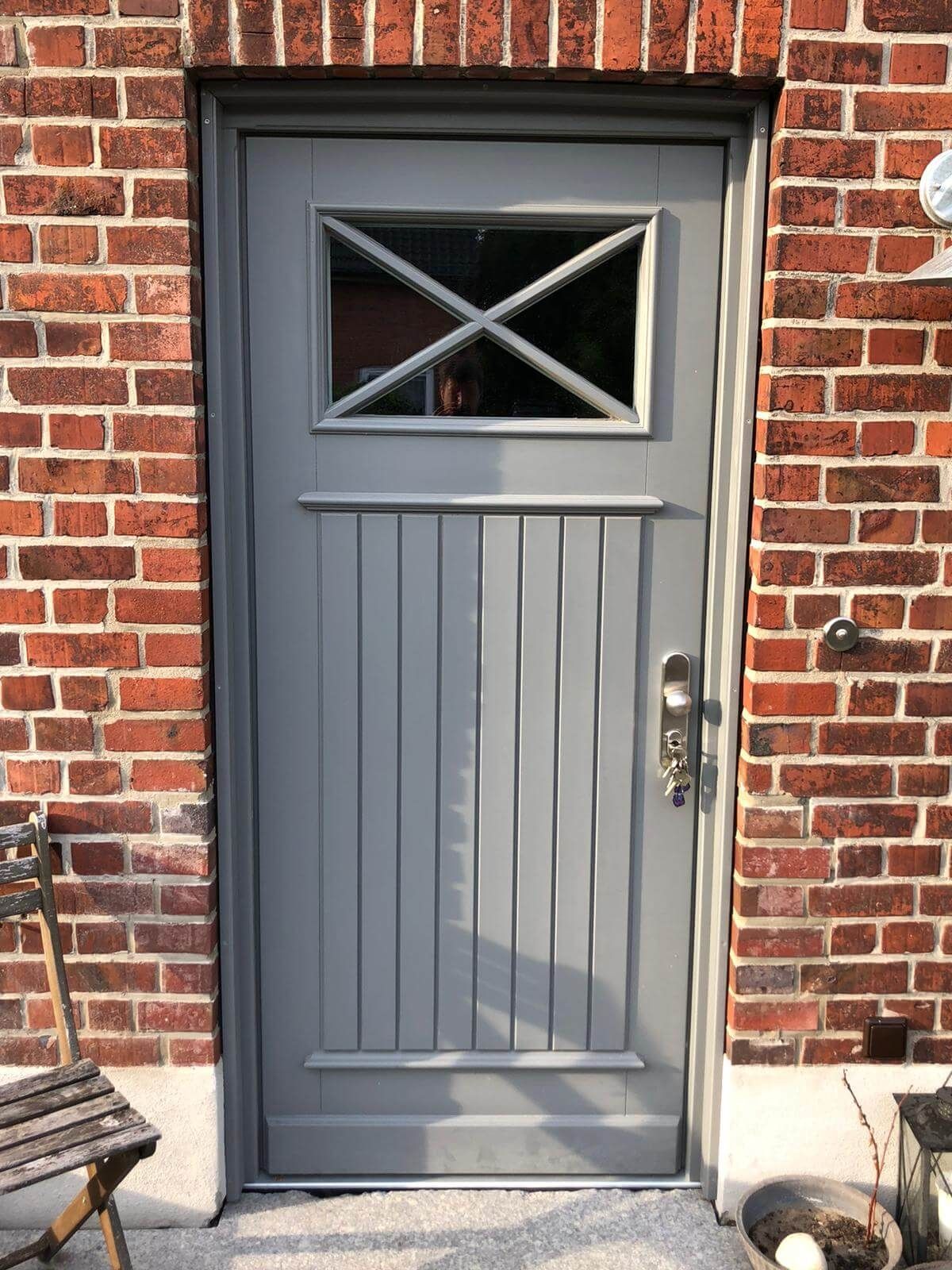 Haustüre im Landhausstil in einer roten Ziegelwand mit rechteckigem Fenster oben. Metallgriff und Schlüssel im Schloss. Links ein Holzstuhl, rechts eine kleine Topfpflanze