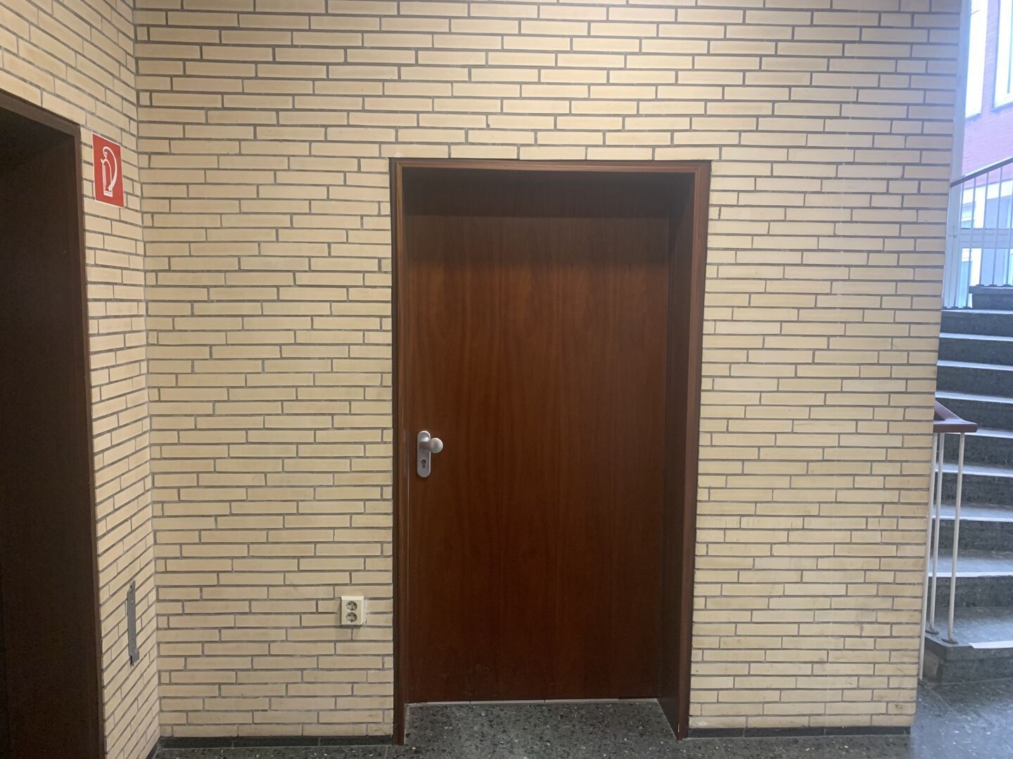 Brandschutztür mit silbernem Griff und rotem Feuerlöscher-Schild daneben. Treppe rechts im Bild