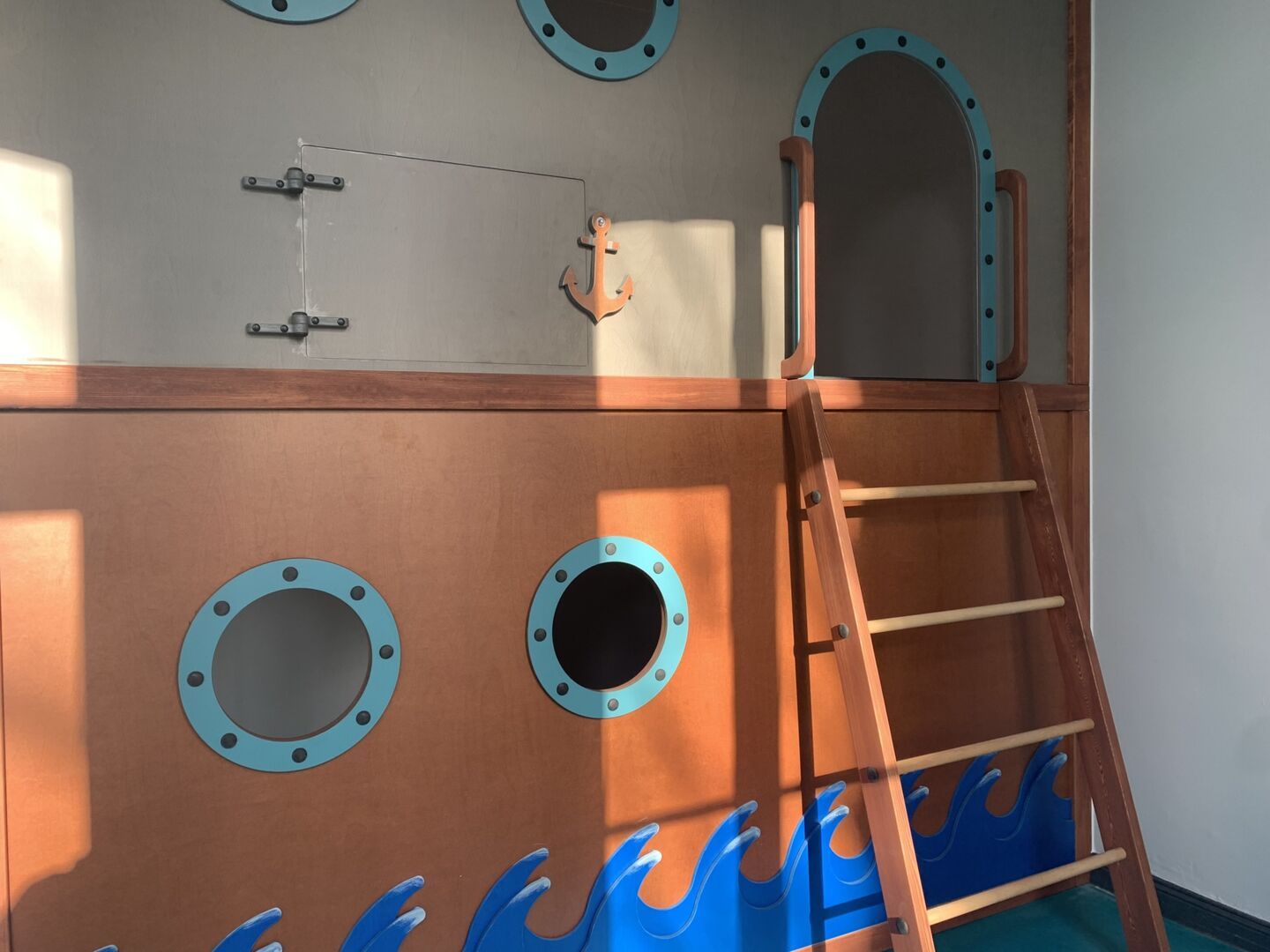 Spielebene in der Kita mit einer Wand in Schiffsoptik, runden Bullaugen-Fenstern und einer Holzleiter, ergänzt durch maritime Details wie eine Anker-Dekoration
