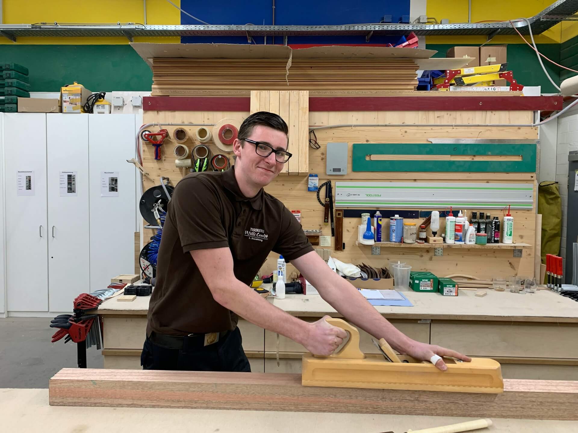 Azubi beim Hobeln eines Holzstücks in einer Werkstatt, umgeben von Werkzeugen und Materialien an einer Rückwand