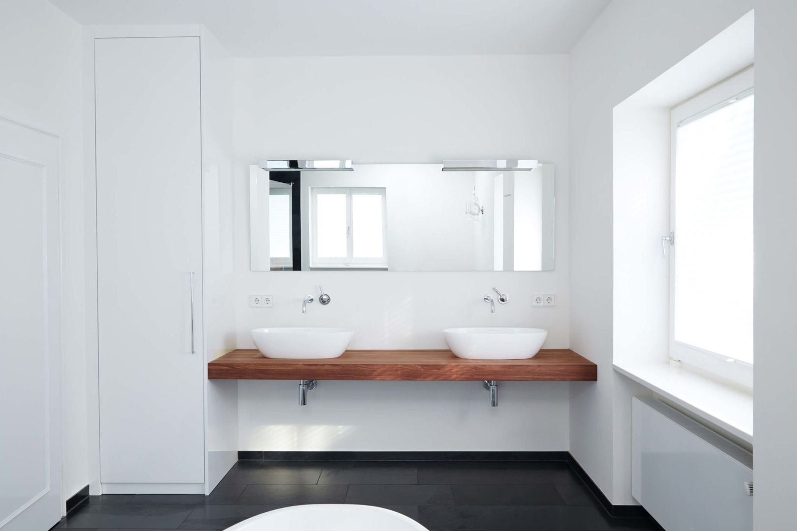 Minimalistisches Badezimmer mit zwei weißen Aufsatzwaschbecken auf einer Holzplatte, großer Spiegel darüber und einem Fenster im Hintergrund