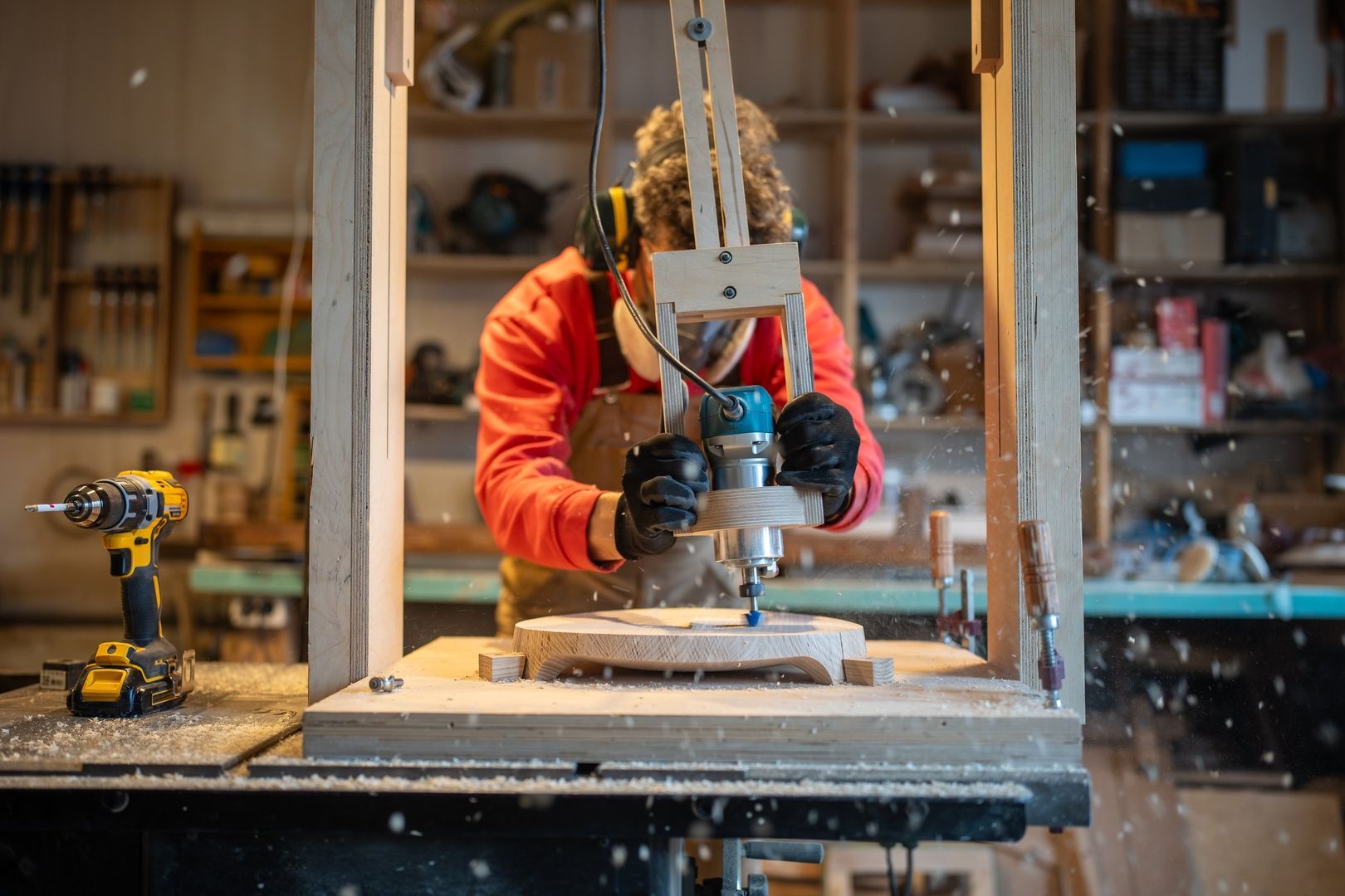 Person arbeitet mit einer Oberfräse in einer Holzwerkstatt, Späne fliegen durch die Luft, im Hintergrund Werkzeuge und Regale
