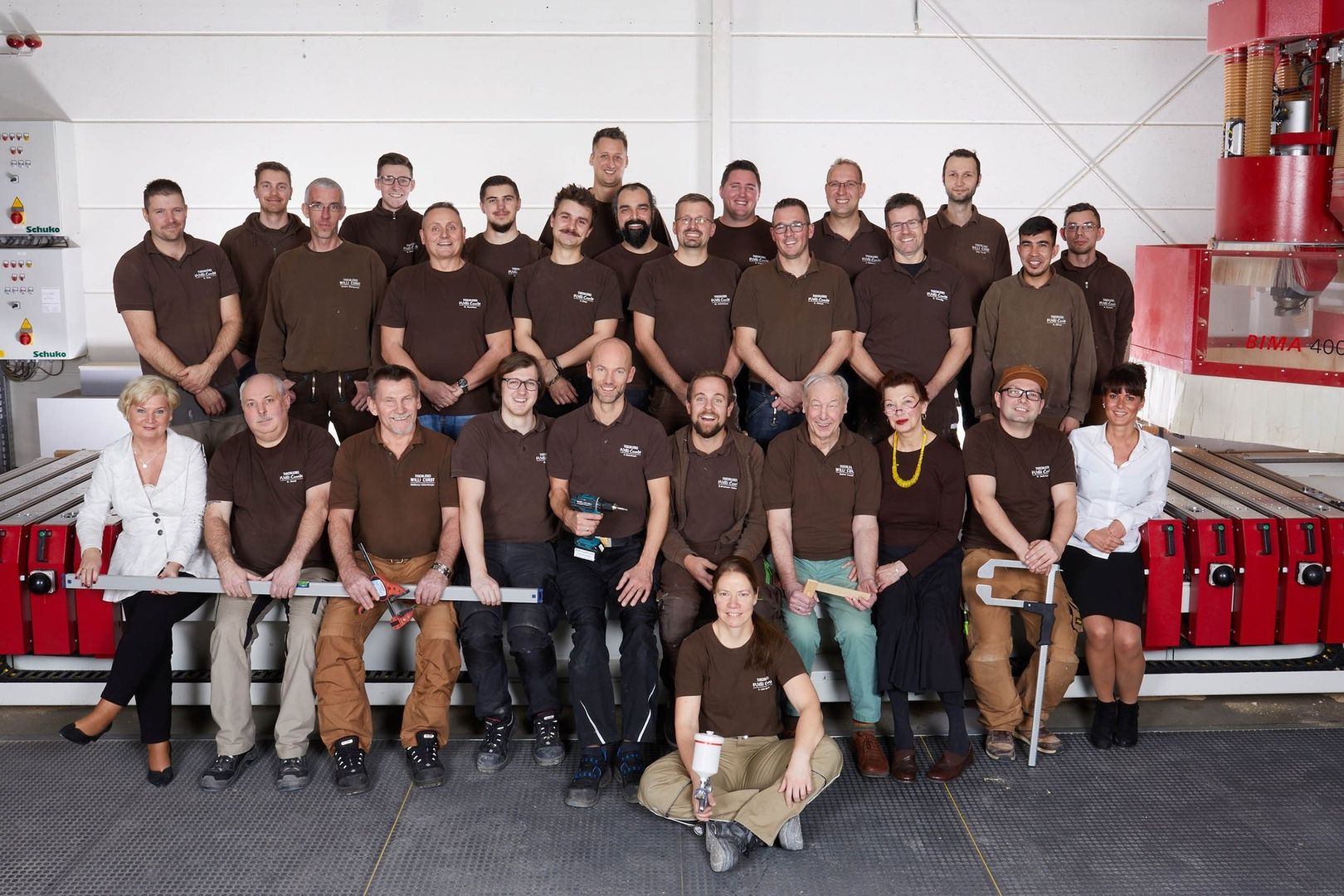 Gruppenbild eines Teams in braunen T-Shirts, aufgenommen in einer Werkstatt mit Maschinen im Hintergrund